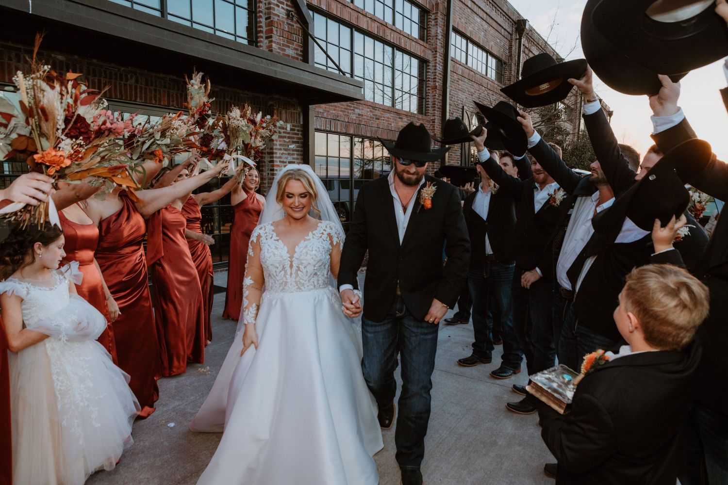 Bride and groom walk through celebratory arch of raised cowboy hats and flowers at rustic wedding exit.