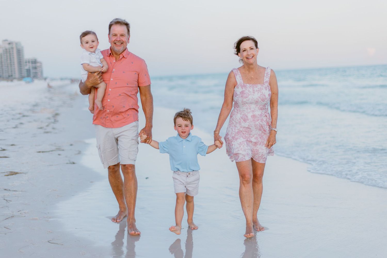 A family wearing coral and white outfits walks together on a sandy beach at sunset.