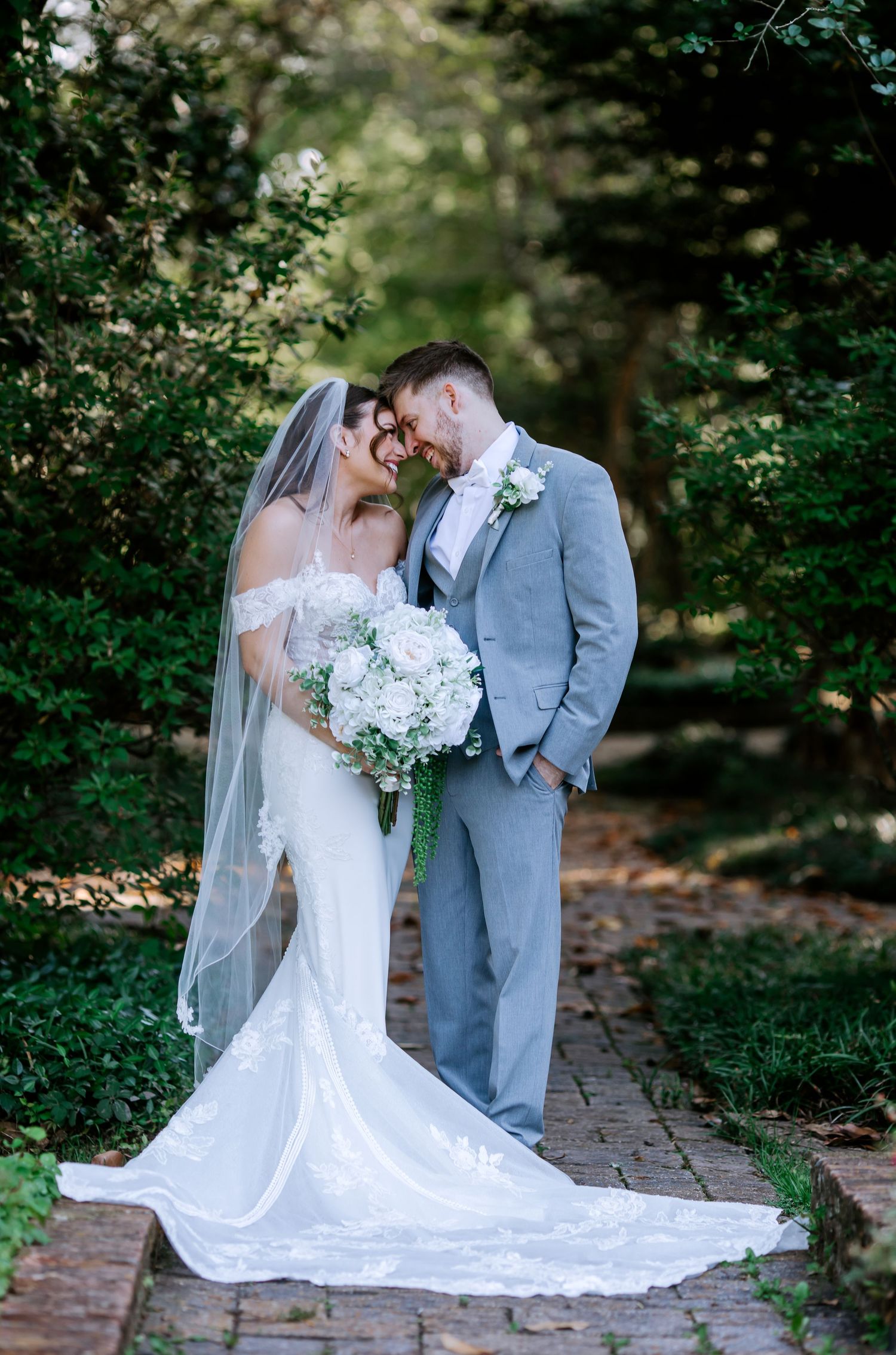 Bride and groom share a romantic forehead touch on a brick path surrounded by lush green trees in an outdoor wedding portrait.
