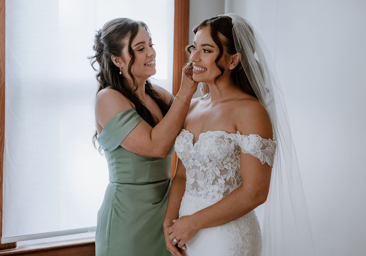 Bridesmaid in sage green dress adjusting bride's veil before wedding ceremony.