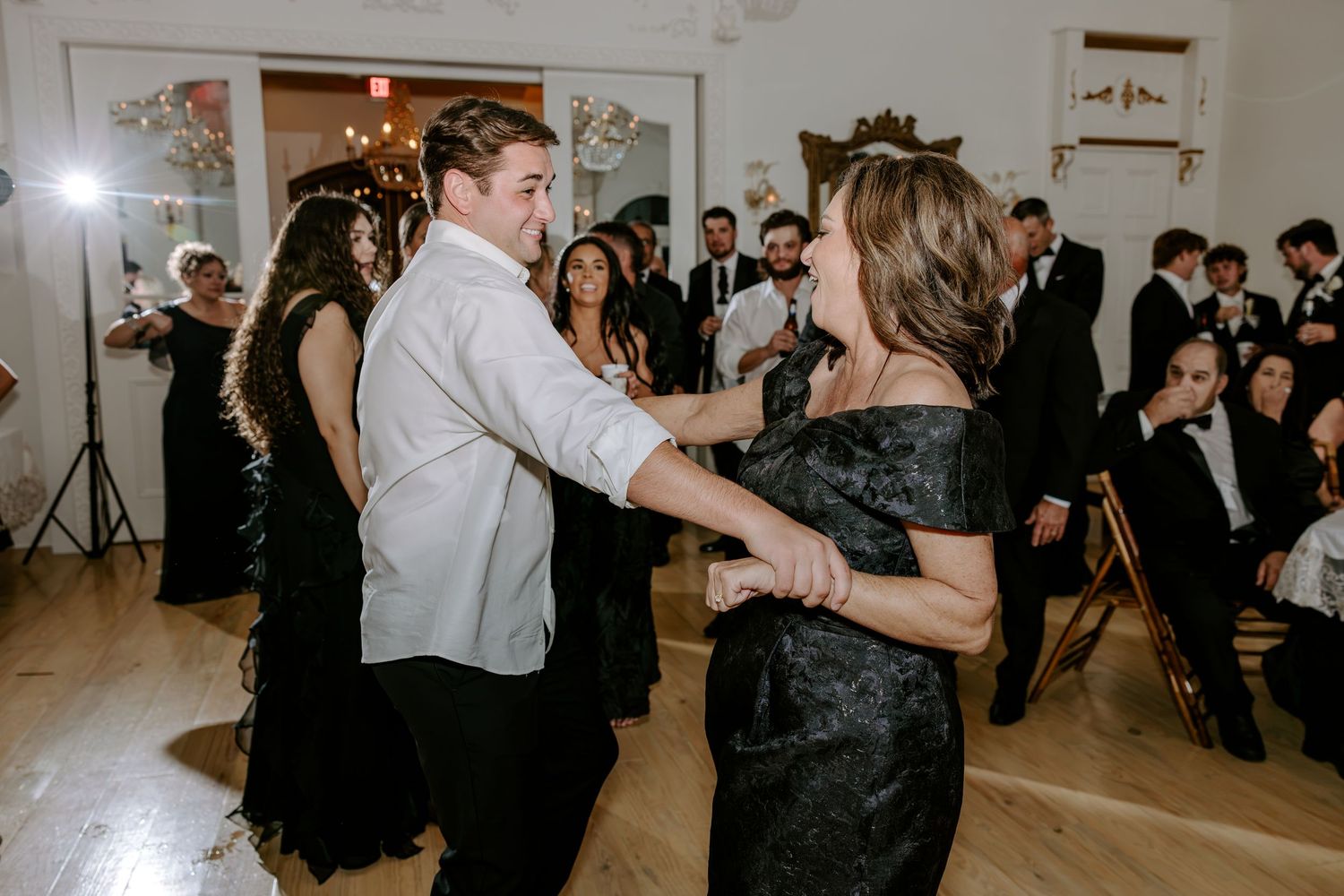 A man and woman in formal attire dance together on a wooden floor while wedding guests watch and smile nearby.