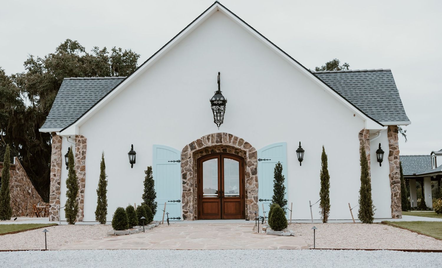 White chapel-style event venue with a stone arched wooden double door, lantern lights, and trimmed evergreen shrubs along a paver walkway.