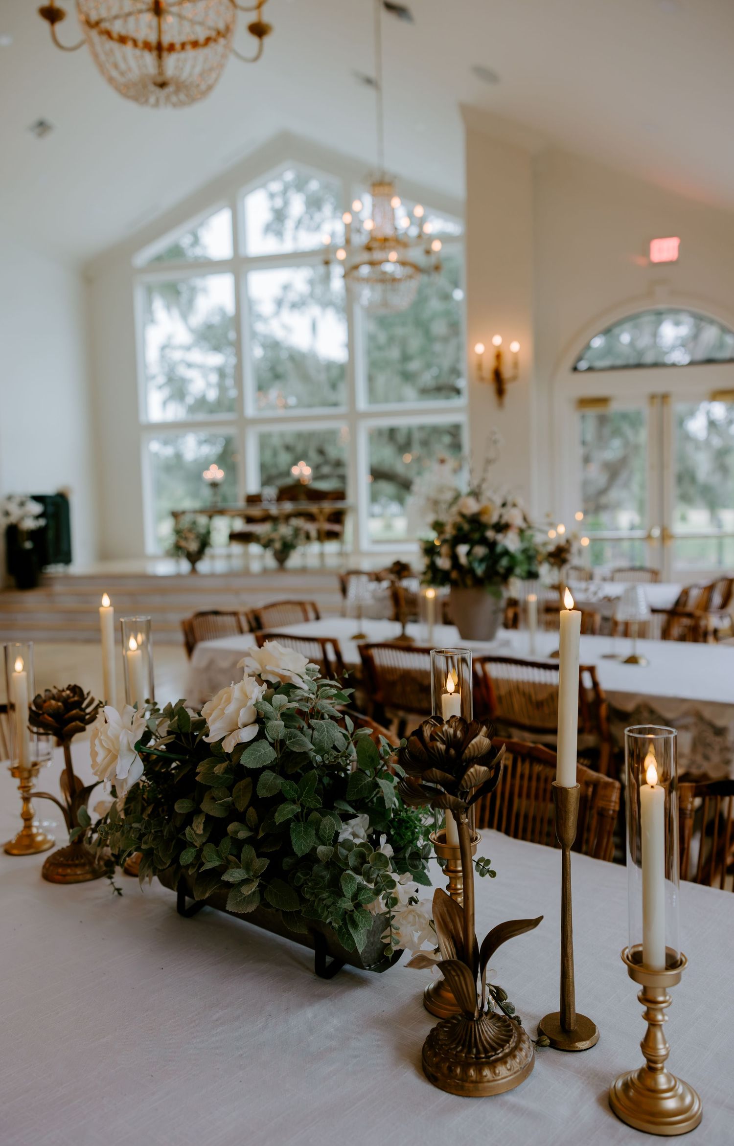 Elegant wedding reception hall with candlelit tables, white floral centerpieces, brass candlesticks, chandeliers, and tall arched windows.