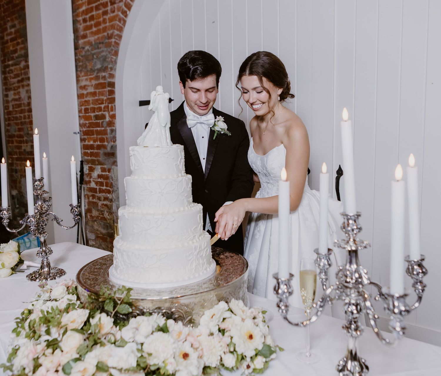 Bride and groom cutting a white four-tier wedding cake surrounded by white flowers and candelabras.