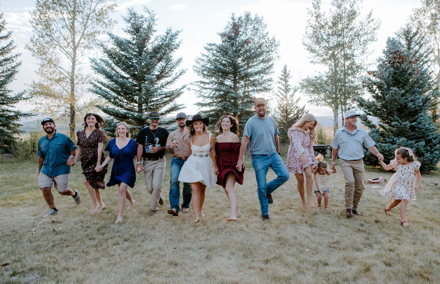 A joyful group of people running and laughing together outdoors near tall evergreen trees on a sunny day.