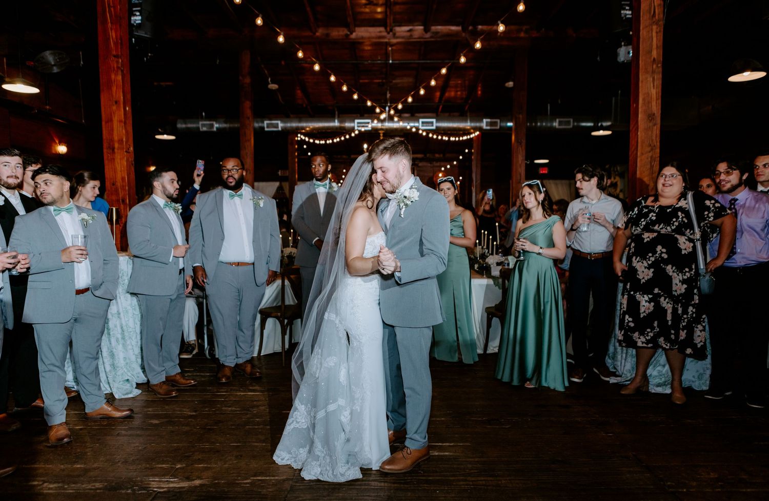 Bride and groom share first dance at rustic wedding reception with guests in teal gowns and gray suits surrounding them.