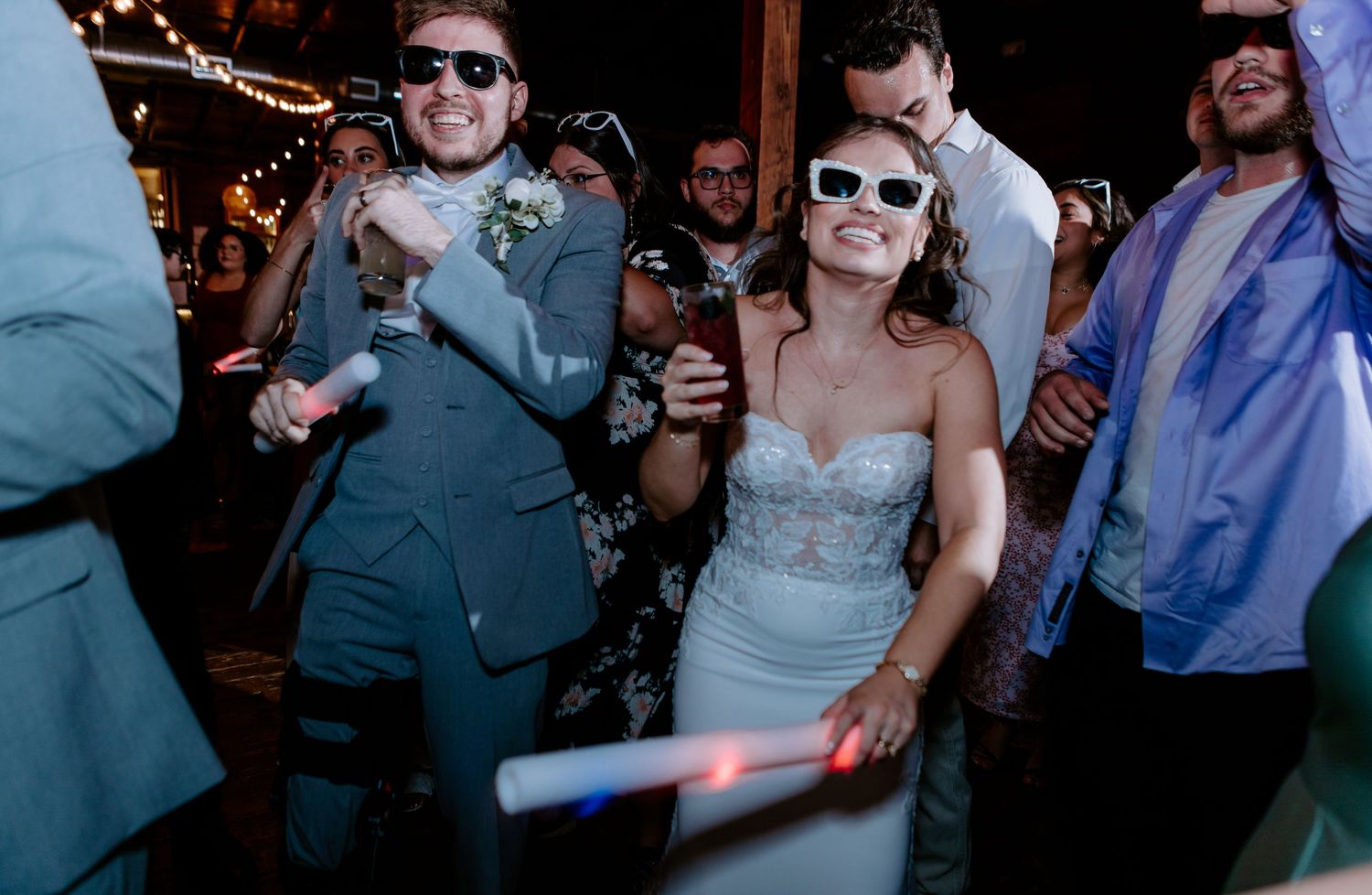 Bride and groom dancing joyfully with guests at wedding reception, wearing fun white sunglasses.