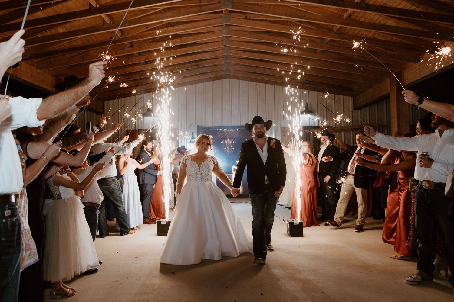 Bride and groom walk through sparkler tunnel held by guests inside a rustic barn wedding venue.