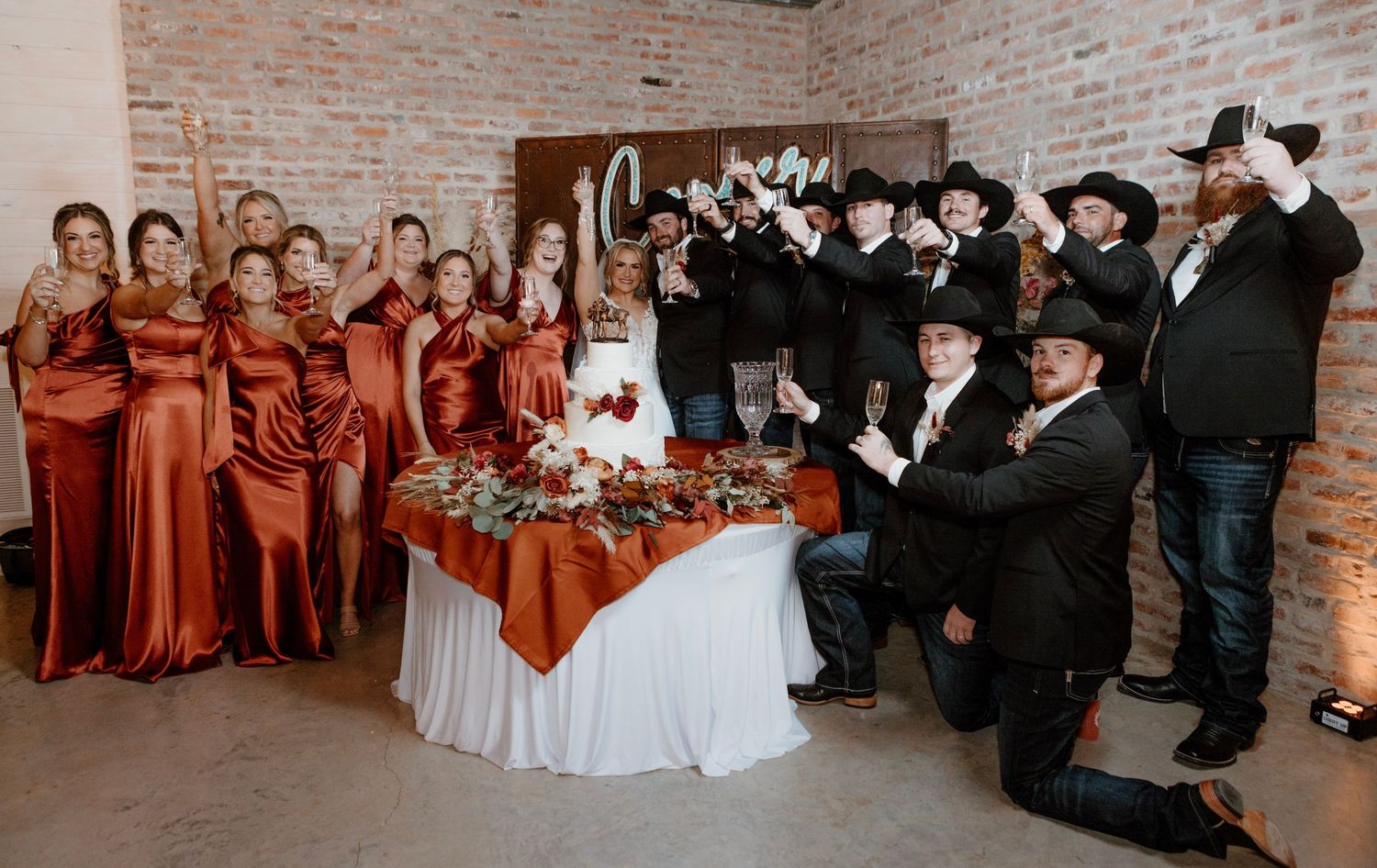 Wedding party in rustic venue, bridesmaids in rust gowns, groomsmen in cowboy hats, couple seated at floral table.