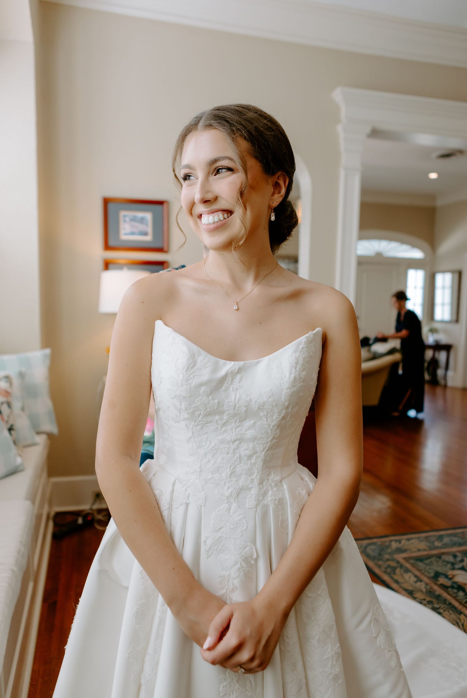 Smiling bride in strapless white lace wedding gown standing in elegant room with hardwood floors.
