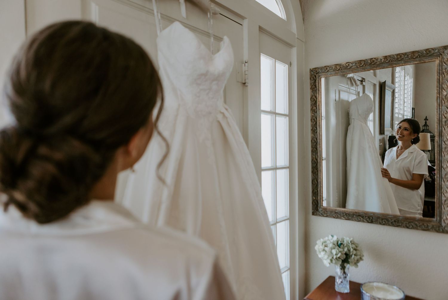 Bride viewing her elegant white wedding gown hanging near a window, reflected in an ornate mirror.