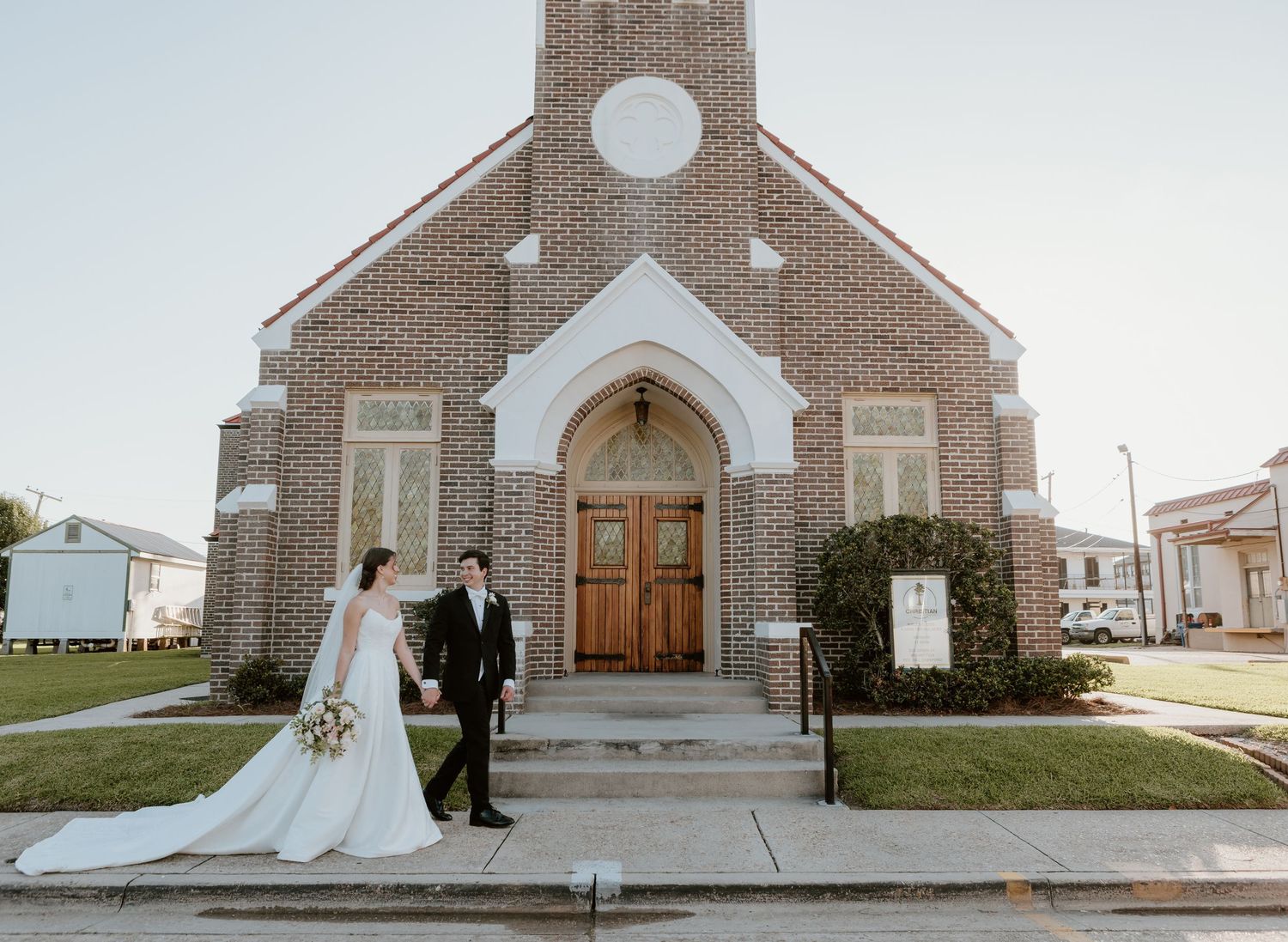 Bride and groom walking hand in hand in front of a brick church with arched wooden doors on their wedding day.