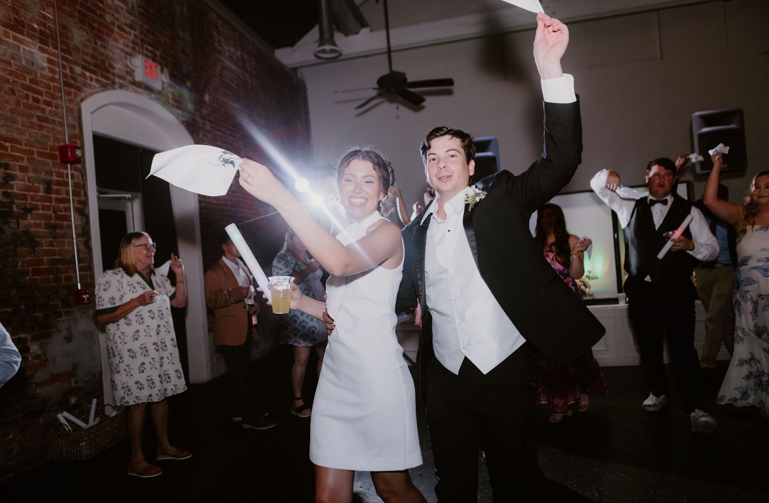 Bride and groom wave napkins joyfully as guests cheer at wedding reception exit.