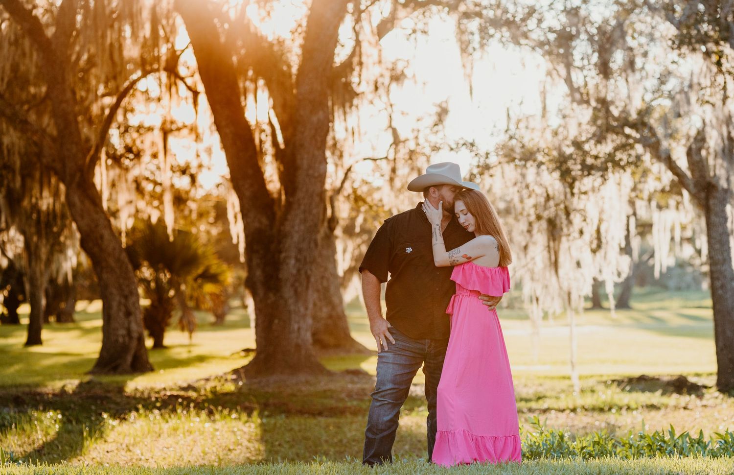 A romantic couple embraces in a sun-drenched park with Spanish moss hanging from oak trees during golden hour.