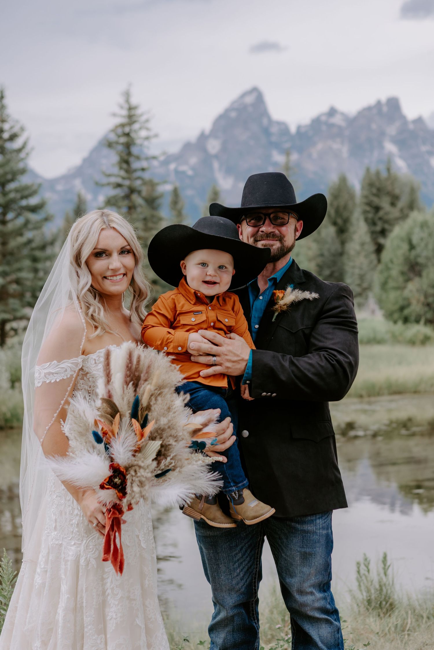 Bride and groom holding toddler in orange shirt pose before Grand Teton mountains at rustic western wedding.