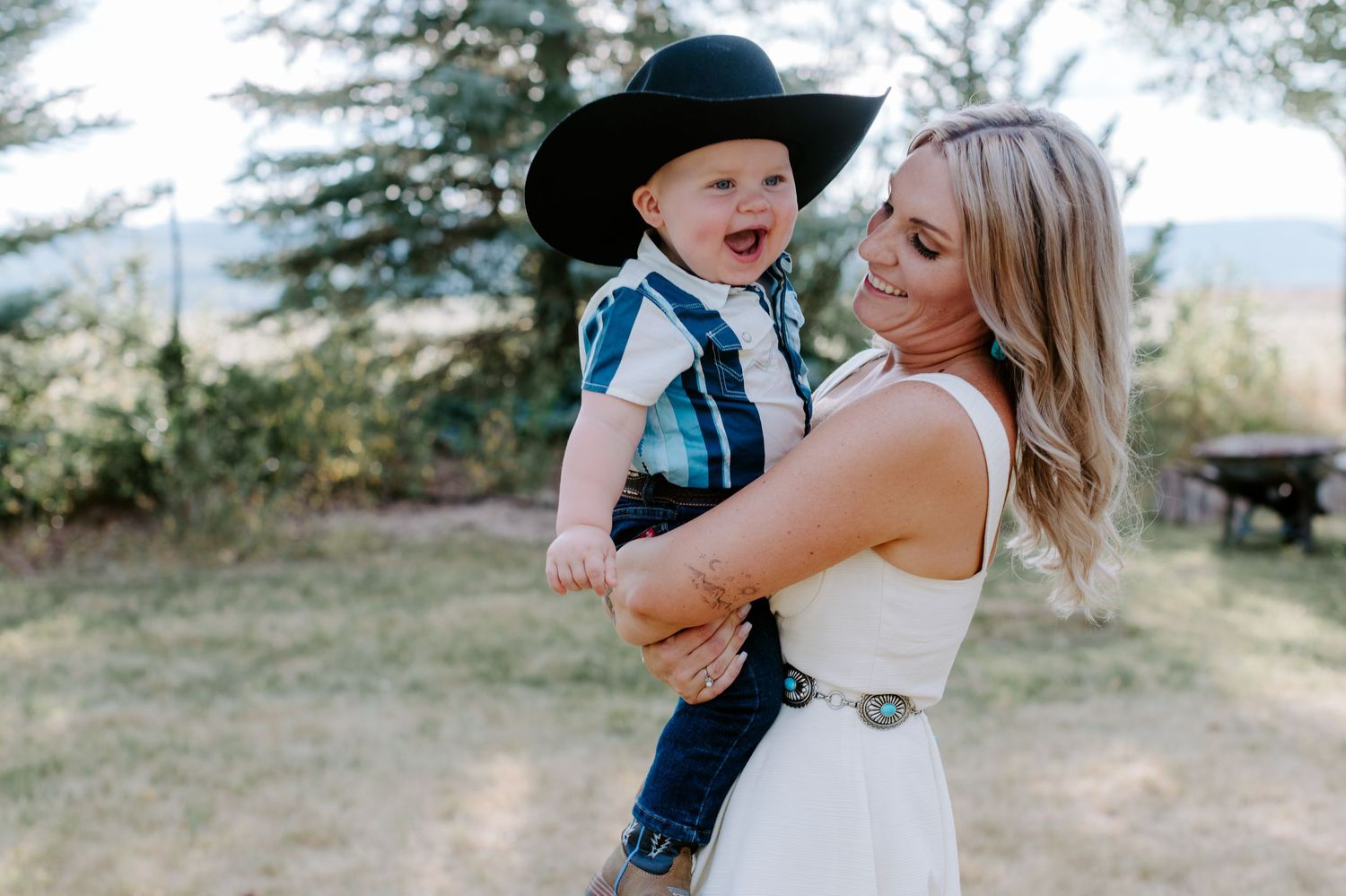 A smiling blonde woman in a white dress holds a laughing toddler boy wearing a black cowboy hat outdoors.
