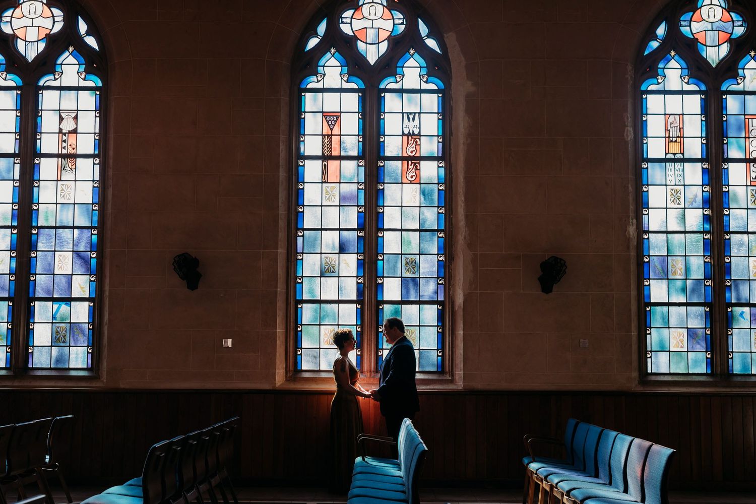 Three tall stained glass windows in a church with blue tones cast light onto wooden pews below.