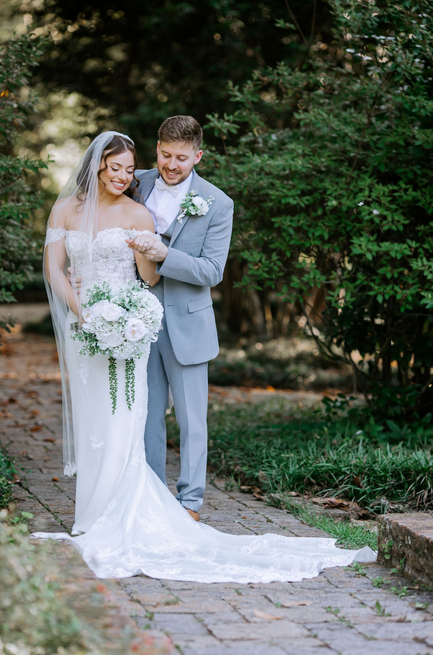 Bride in strapless lace gown with cathedral veil and groom in gray suit share a tender moment on brick pathway.
