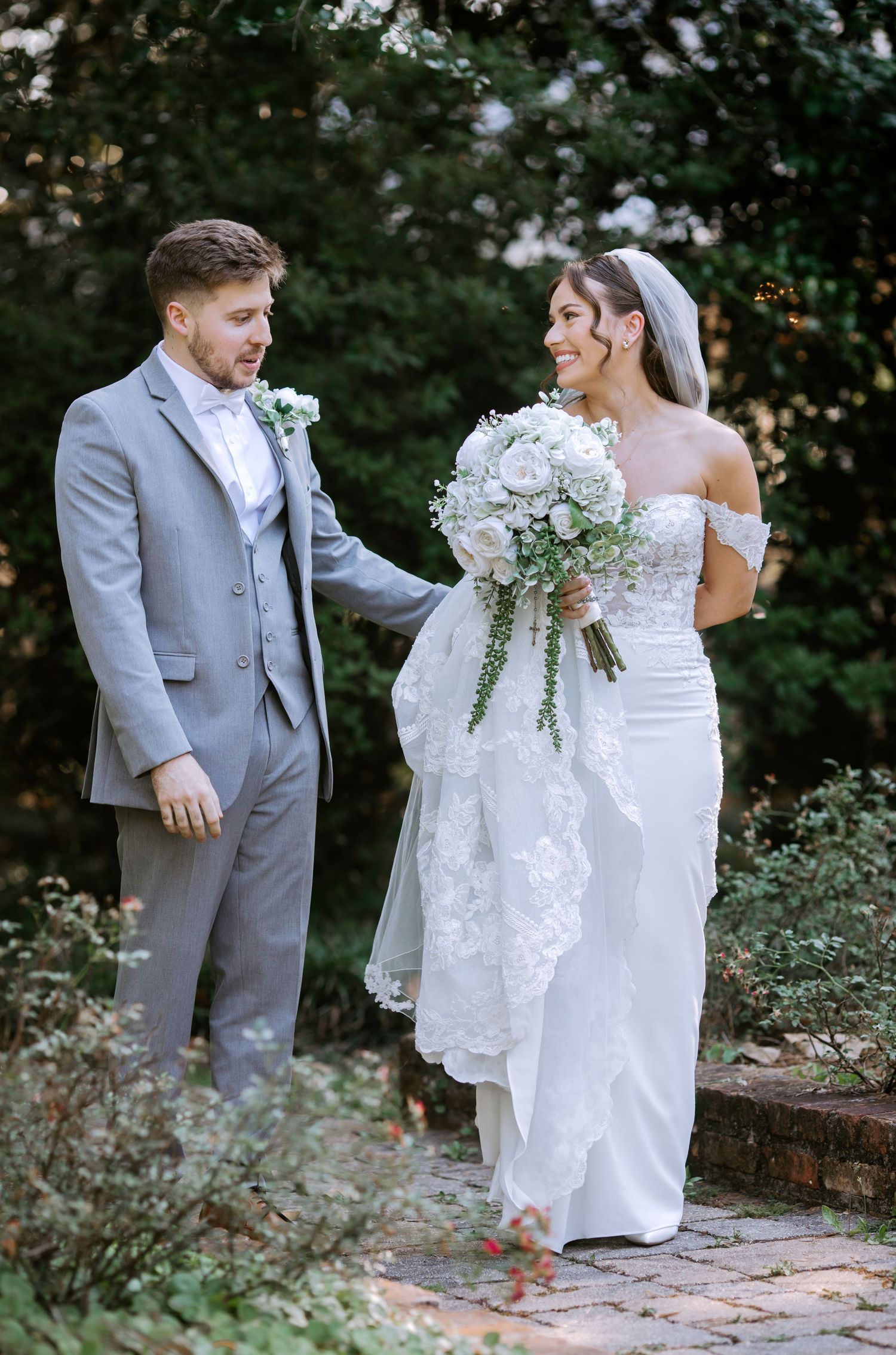 Groom in gray suit holds bride's lace train as she smiles holding white roses on a brick garden path.