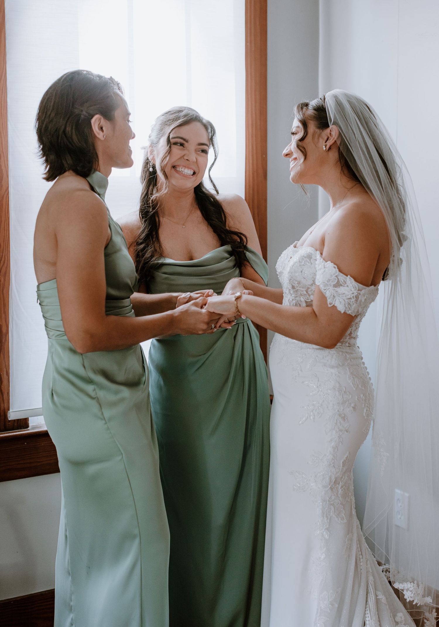 Bride in lace gown with two bridesmaids in sage green dresses sharing a joyful moment before the wedding ceremony.