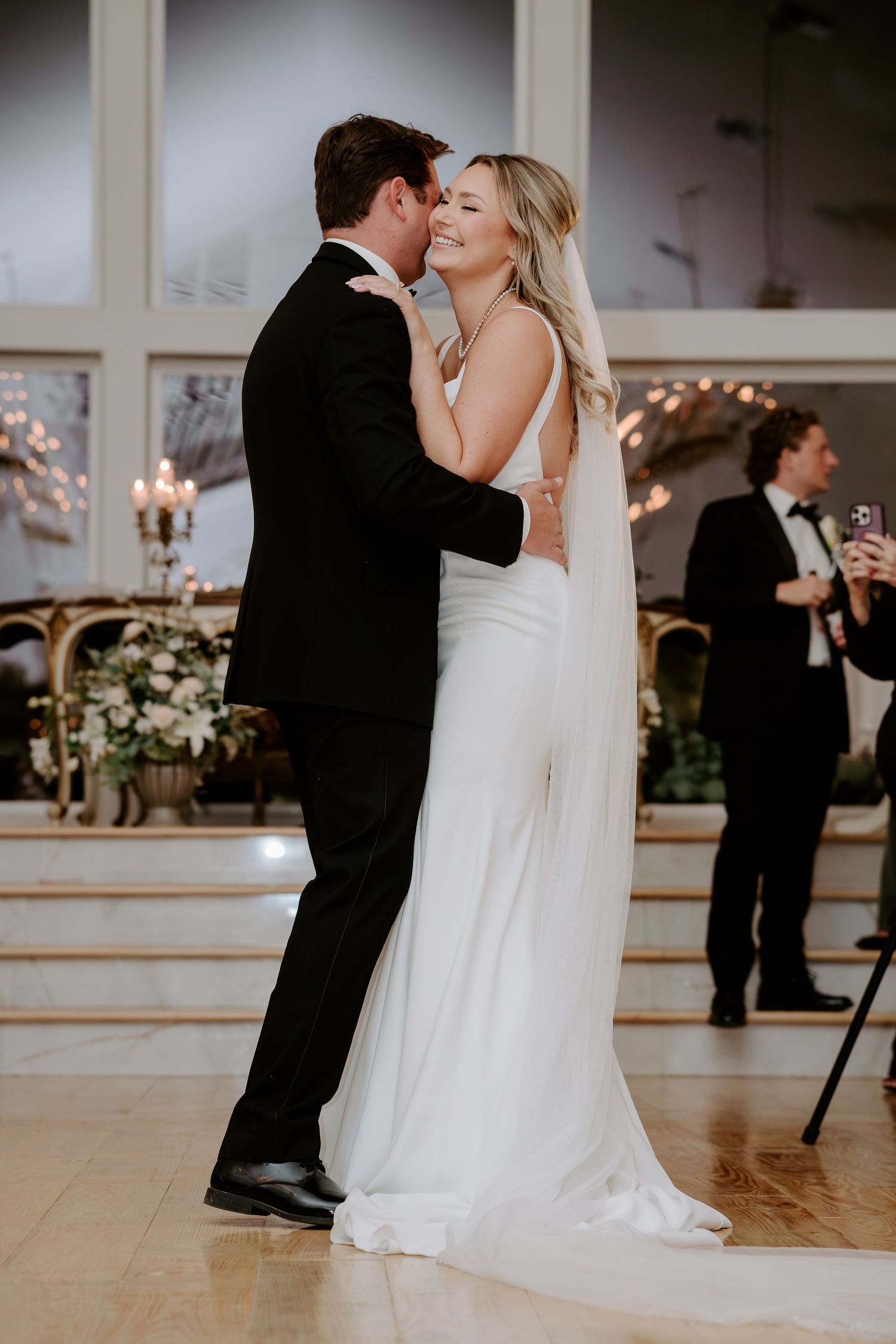 Smiling bride in a white gown and veil dances closely with a groom in a black suit during their first dance at an elegant wedding reception.