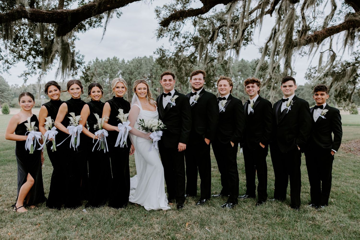 Bride in a white gown holding a bouquet stands with bridesmaids in black dresses and groomsmen in tuxedos under mossy oak trees.