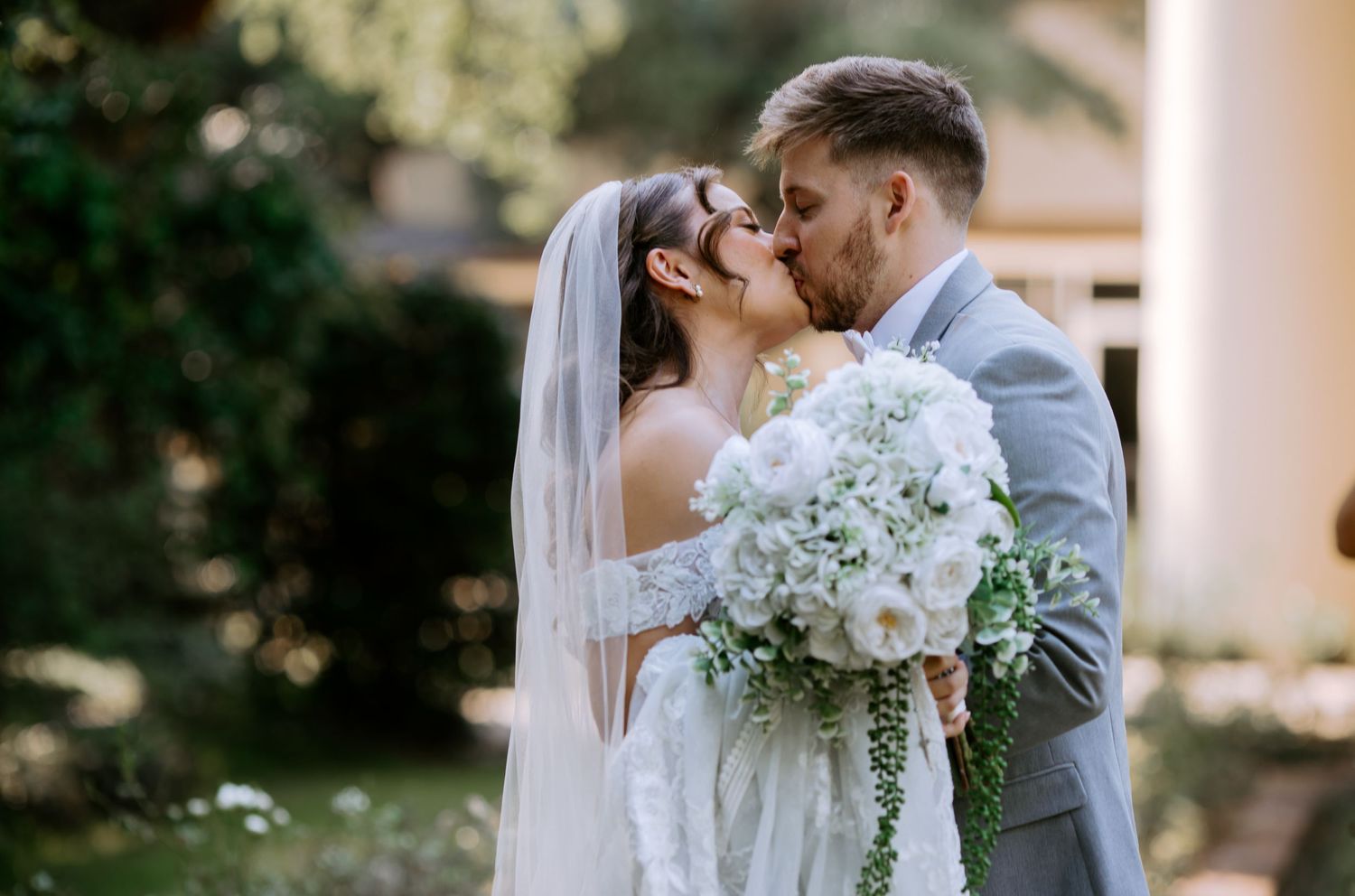 Bride and groom share a romantic kiss outdoors, bride holding white floral bouquet wearing veil and lace gown.