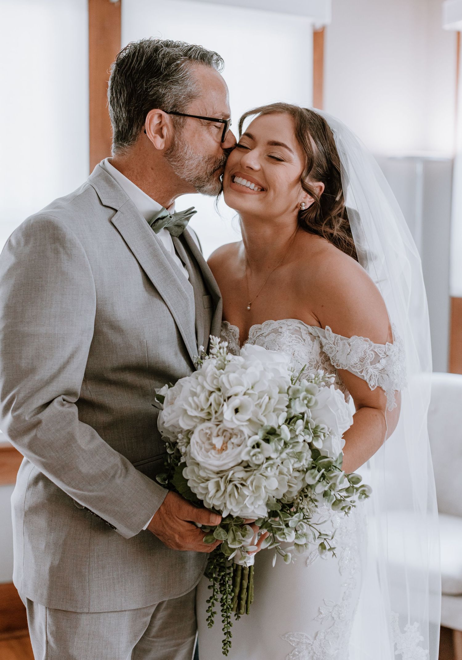 Groom in gray suit kissing bride wearing off-shoulder lace gown holding white hydrangea bouquet on wedding day.