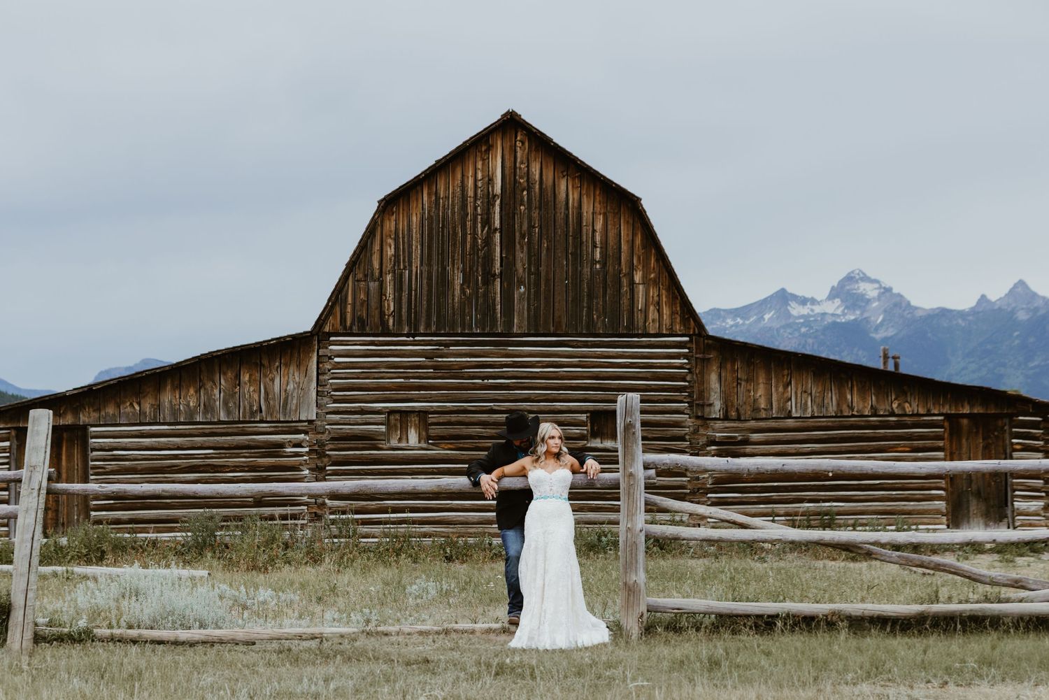 Bride and groom pose before a rustic wooden barn with Grand Teton mountains in background at Jackson Hole wedding.
