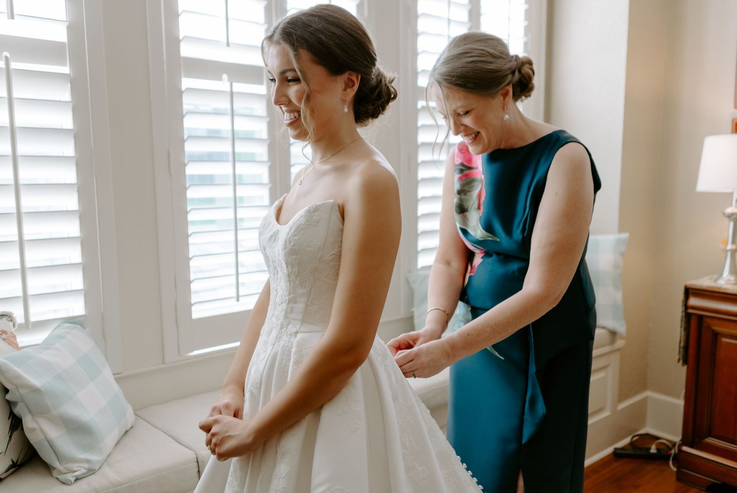 Bride in white gown getting dress fastened by woman in teal dress near bright window.