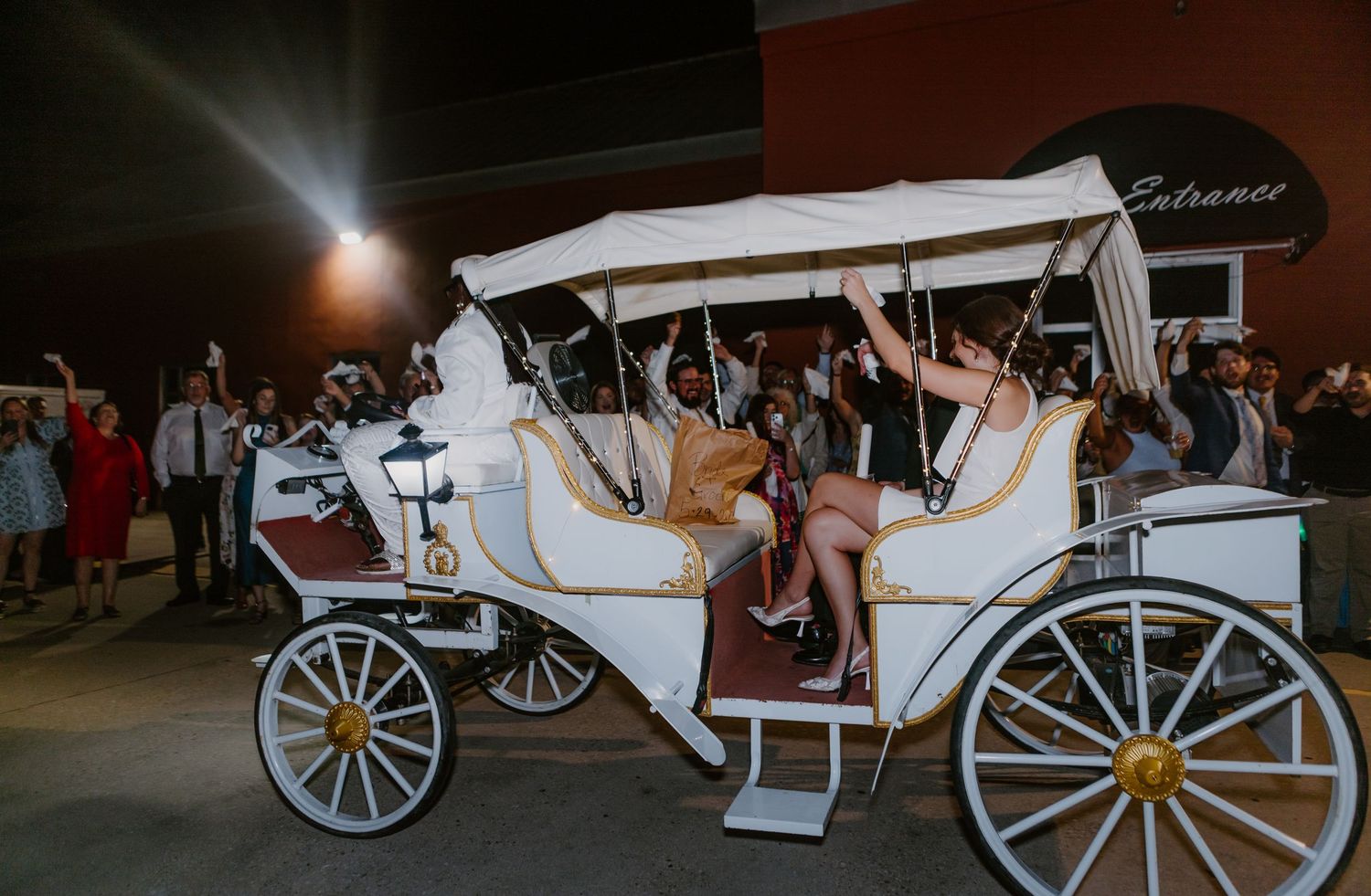 Wedding couple waves from elegant white horse-drawn carriage surrounded by celebrating guests.
