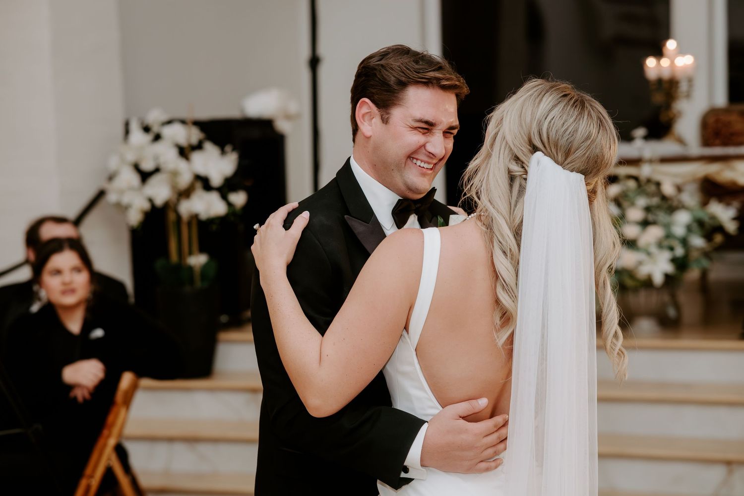 Smiling groom in a black tuxedo shares a first dance with his bride in a white gown and long veil, surrounded by white flowers and wedding g