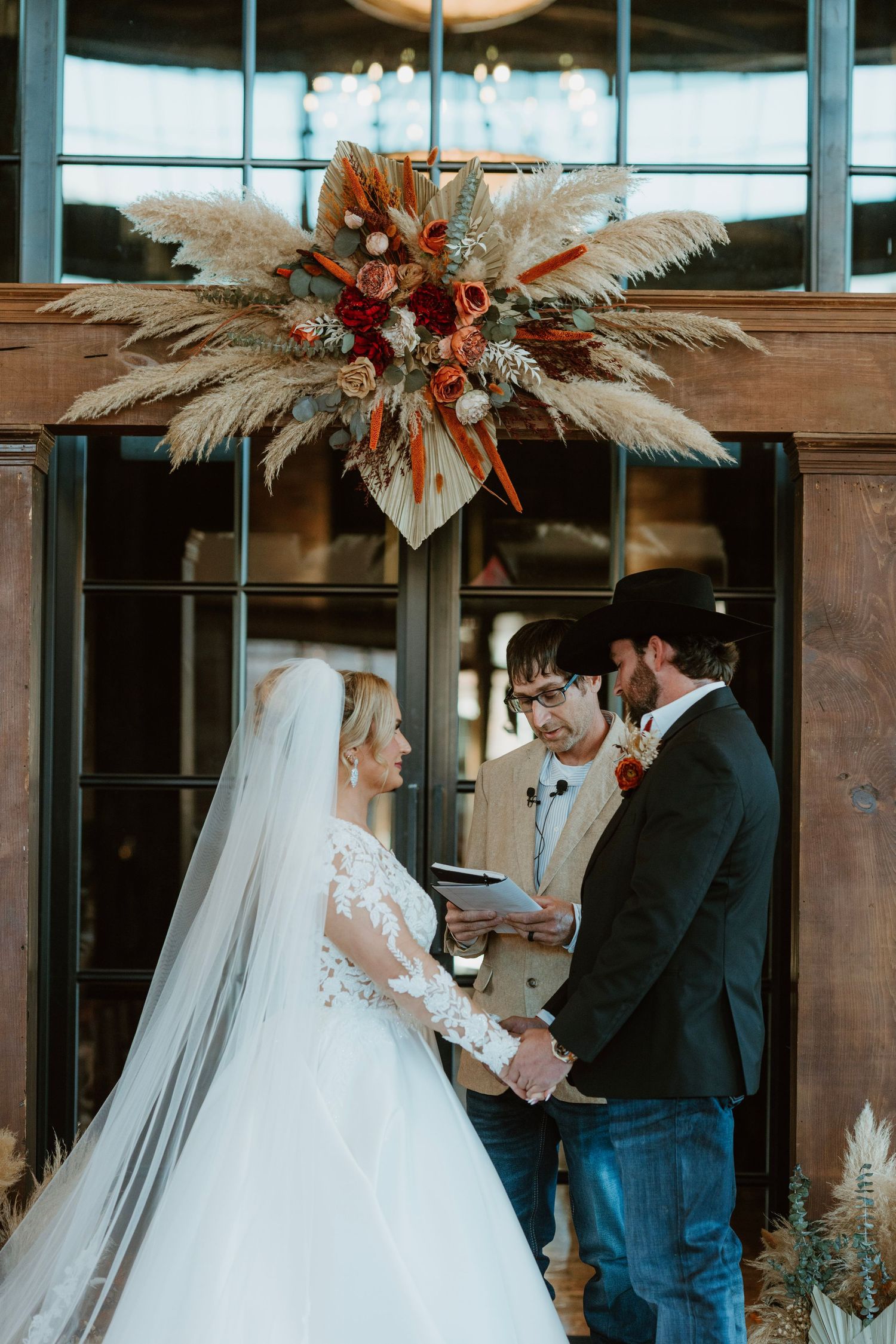 Bride and groom exchange vows beneath a boho pampas grass floral arch at a rustic indoor wedding ceremony.