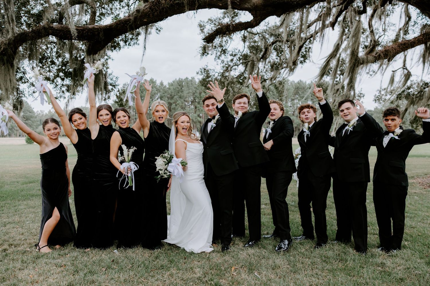 Bride in white gown surrounded by cheering bridesmaids and groomsmen in black attire under oak trees draped with Spanish moss.