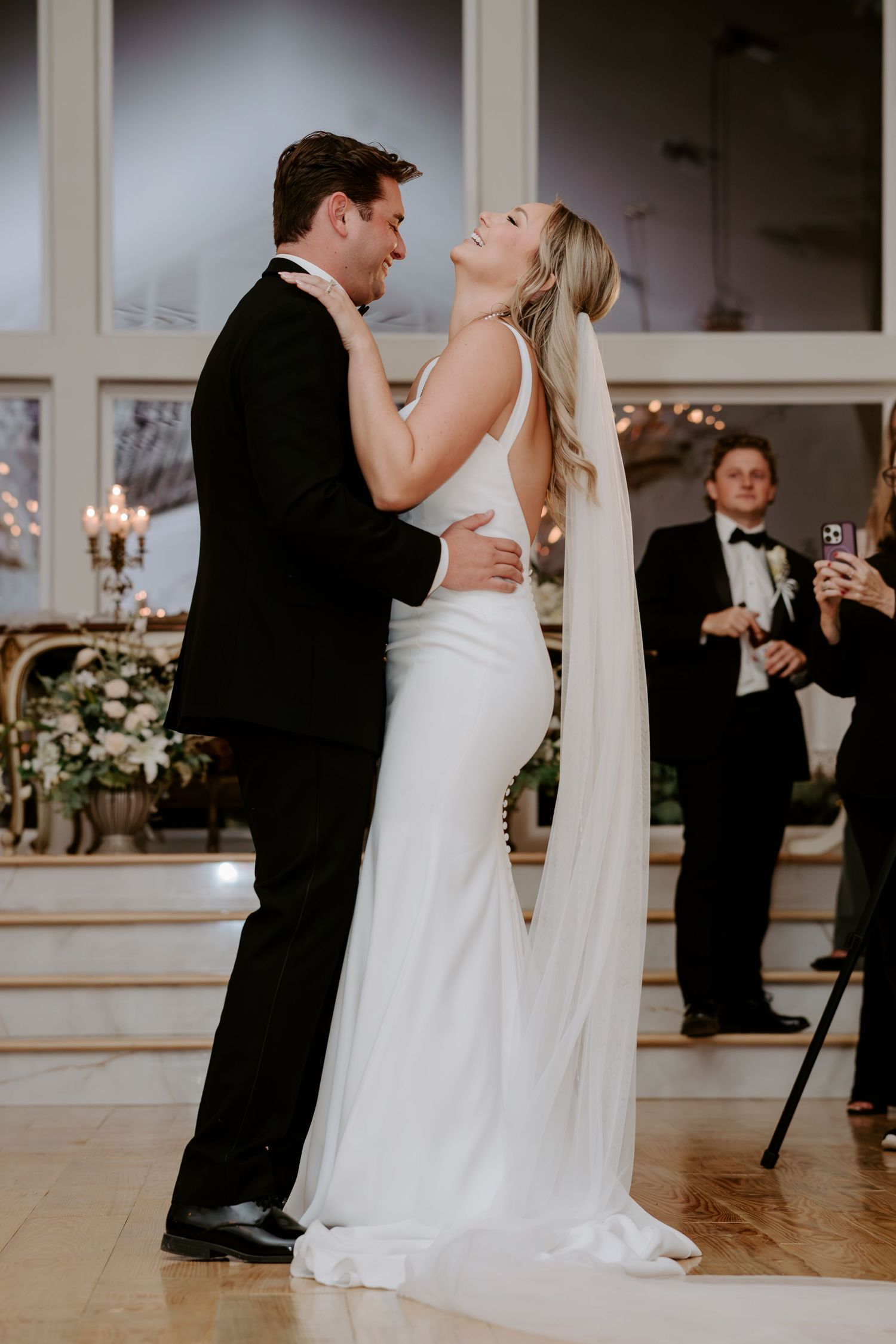 Bride in a fitted white gown and long veil laughs joyfully while dancing with her groom in a black tuxedo at an elegant wedding reception.