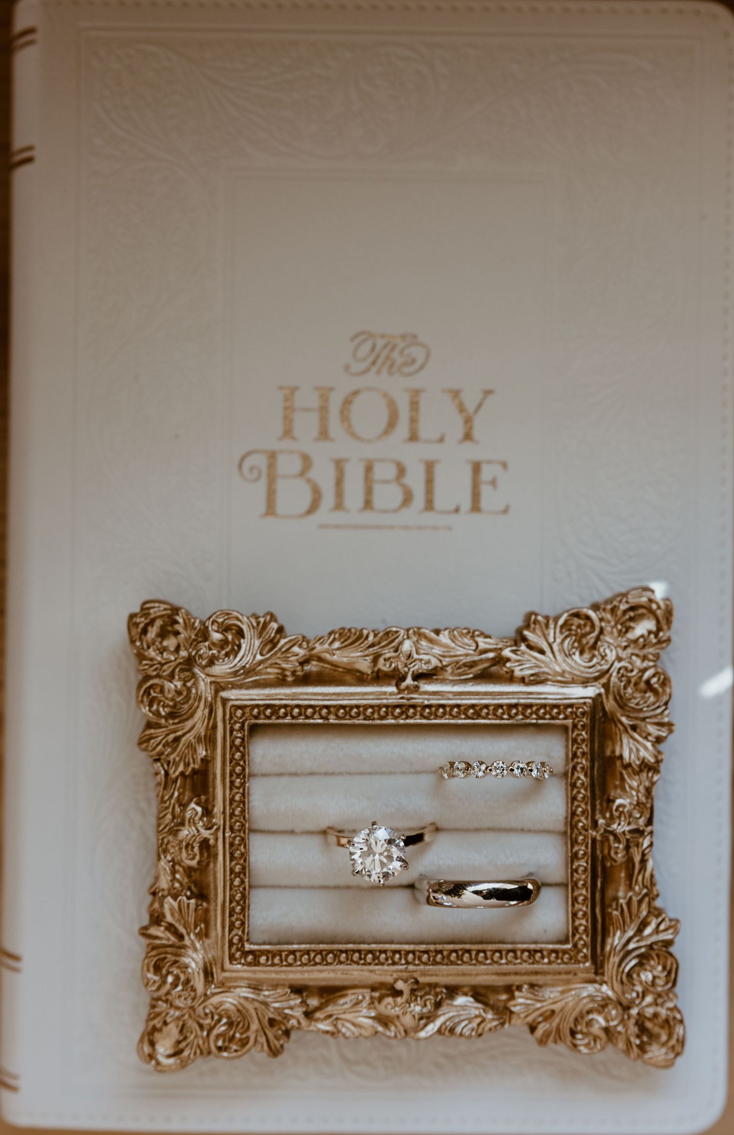 Wedding rings, including a diamond engagement ring and two bands, displayed in an ornate gold frame on a cream Holy Bible.