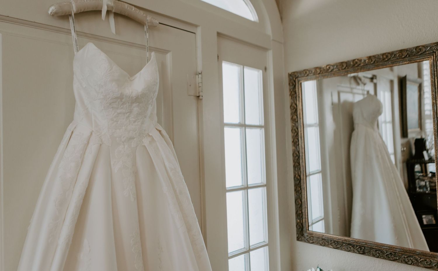 Elegant white wedding dress hanging near a mirror in a bright bridal suite.