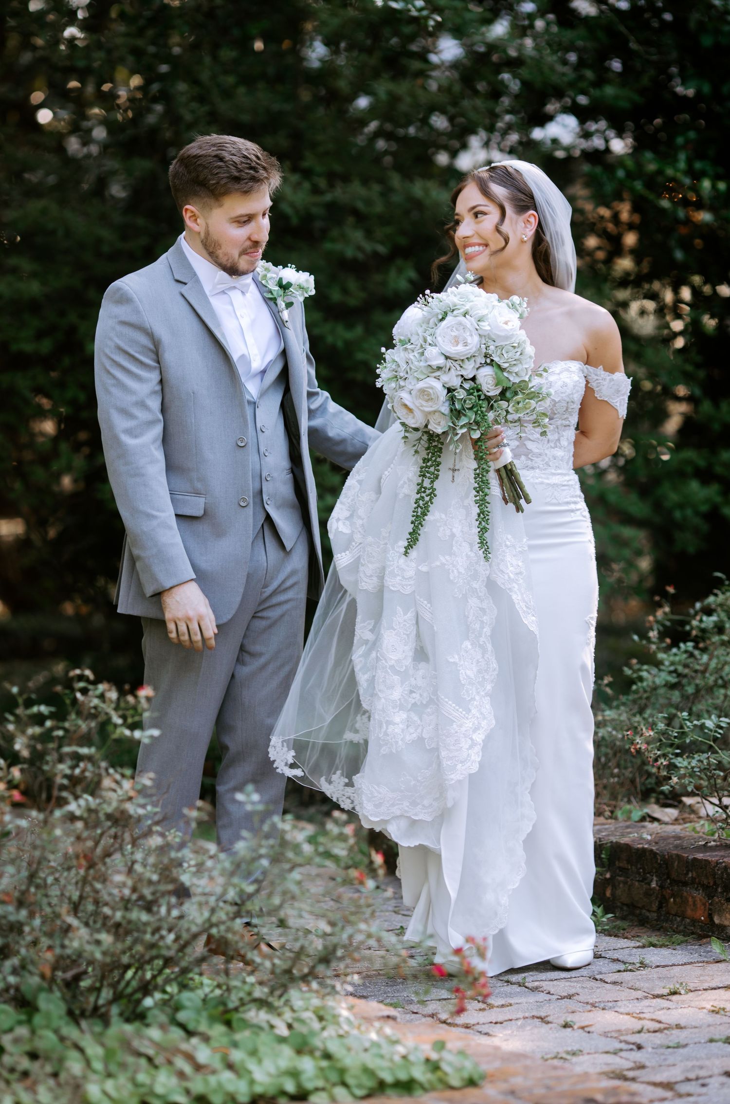 Newlyweds walk together in garden, bride in off-shoulder lace gown carrying white floral bouquet with greenery.