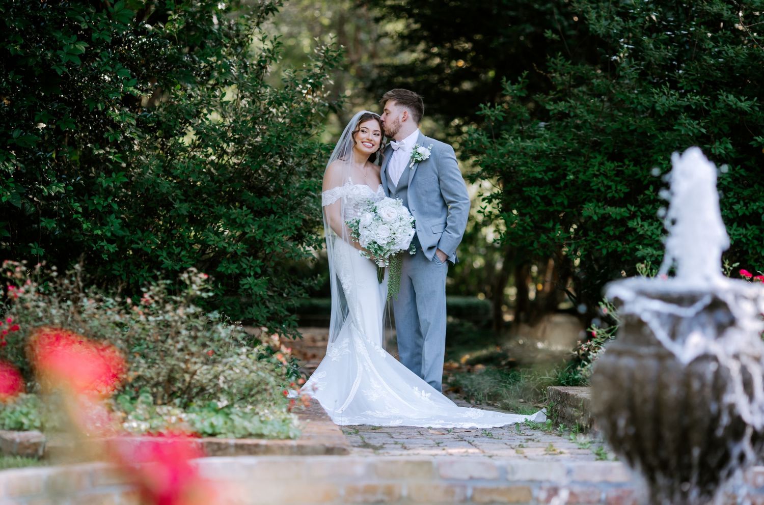 Bride and groom sharing a kiss near a garden fountain, bride in elegant white gown with long veil and white bouquet.