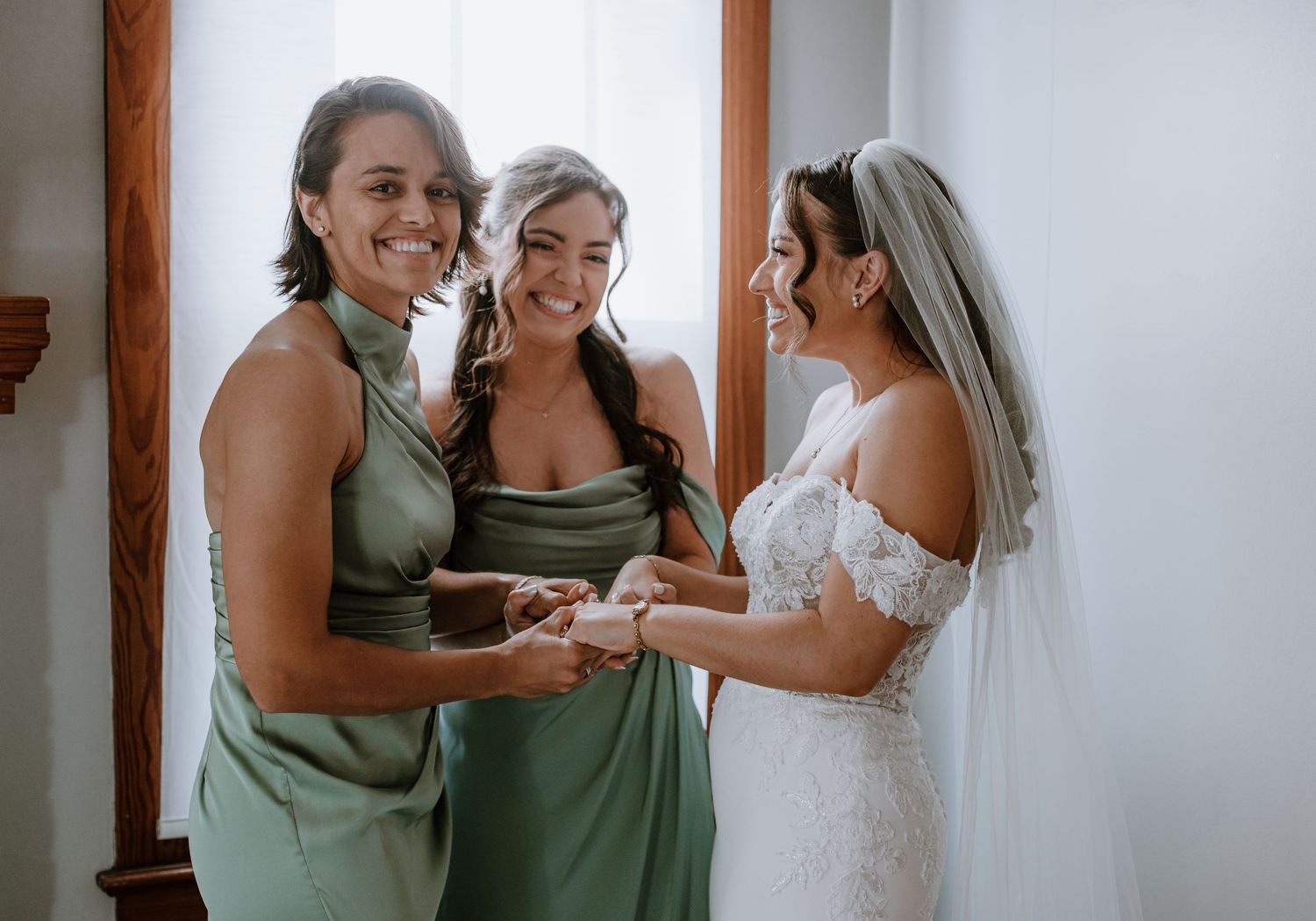 Bride in lace gown and veil shares a joyful moment with two bridesmaids in sage green dresses before the wedding ceremony.