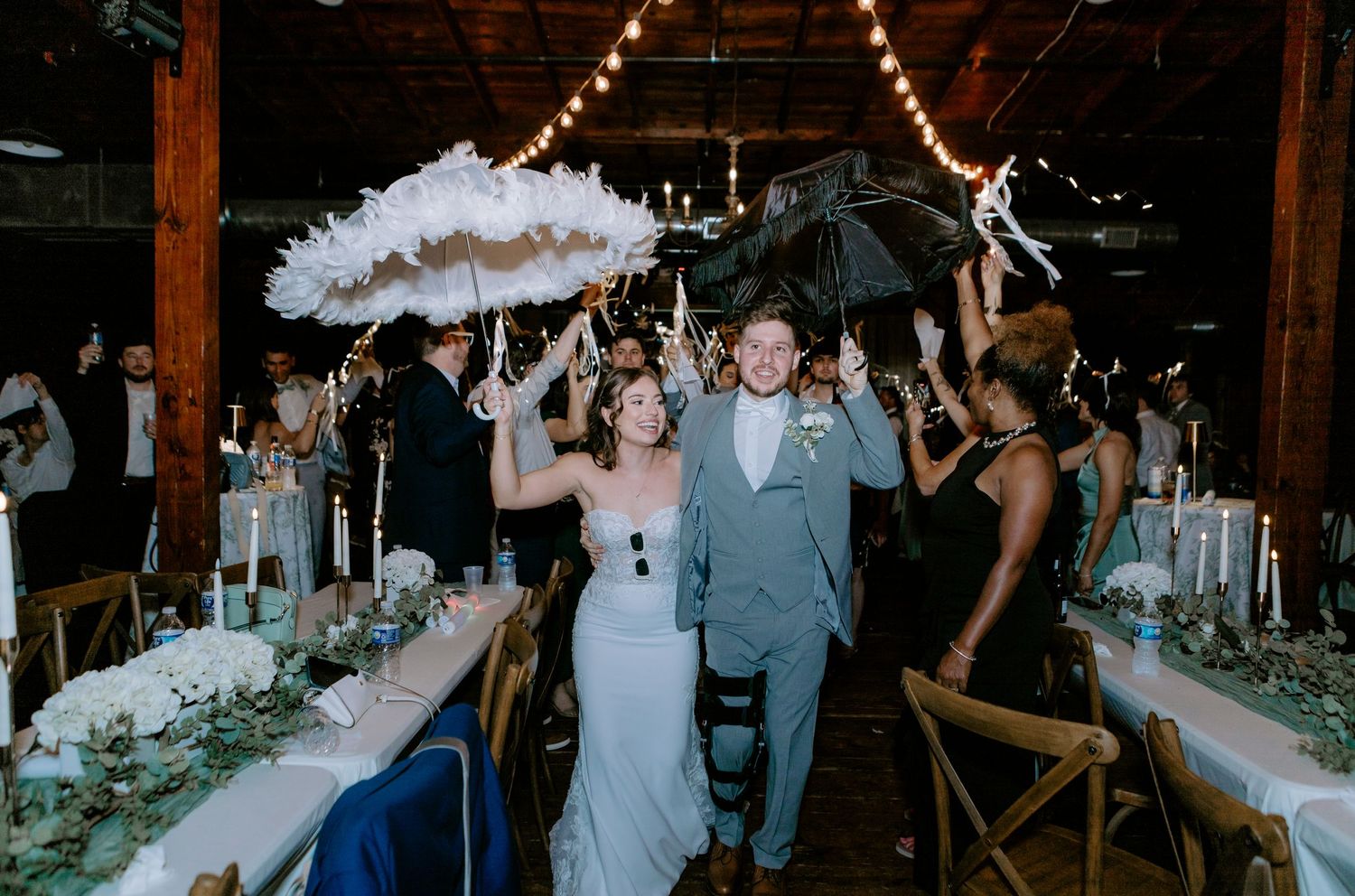 Bride and groom walk through reception hall as guests celebrate with feather fans and umbrellas at elegant wedding.