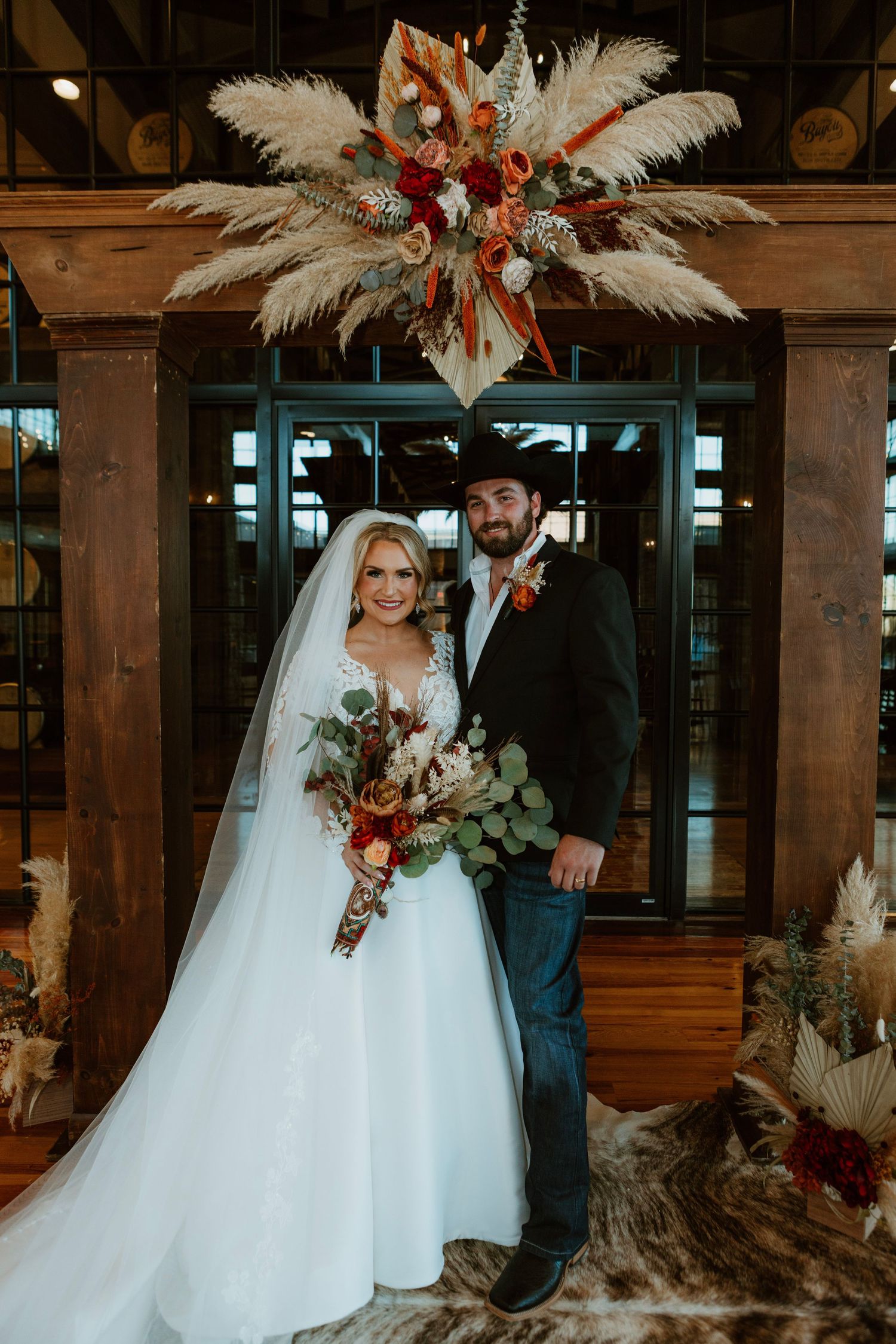 Bride in white lace gown and groom in black suit pose under boho floral arch with pampas grass at rustic wedding venue.