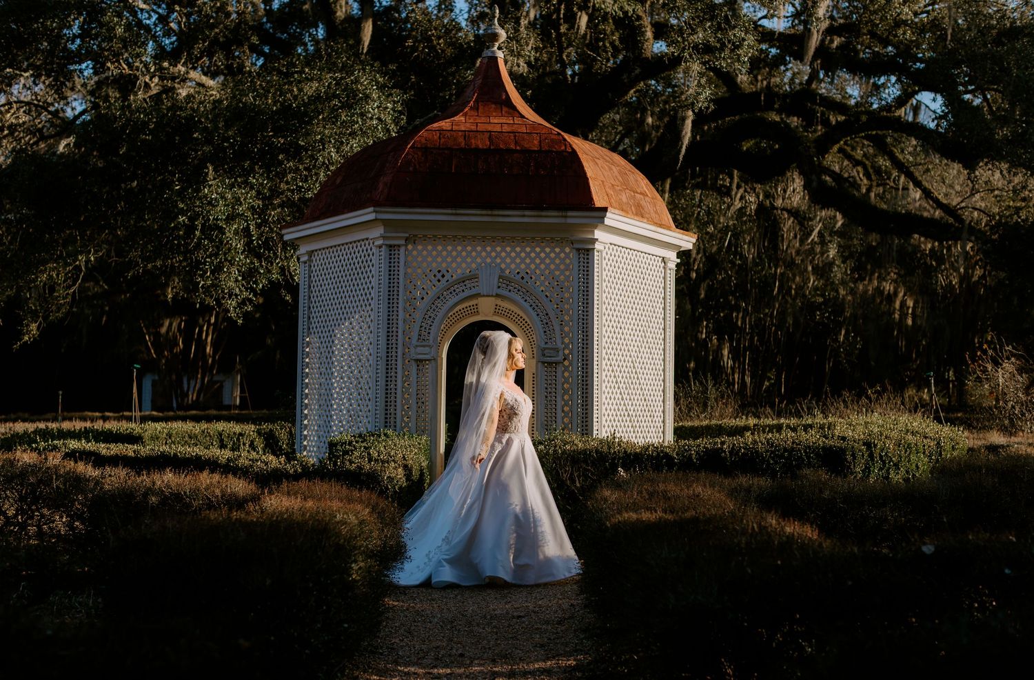 A romantic outdoor wedding photoshoot with a gazebo and flowing white dress in a garden setting at sunset.