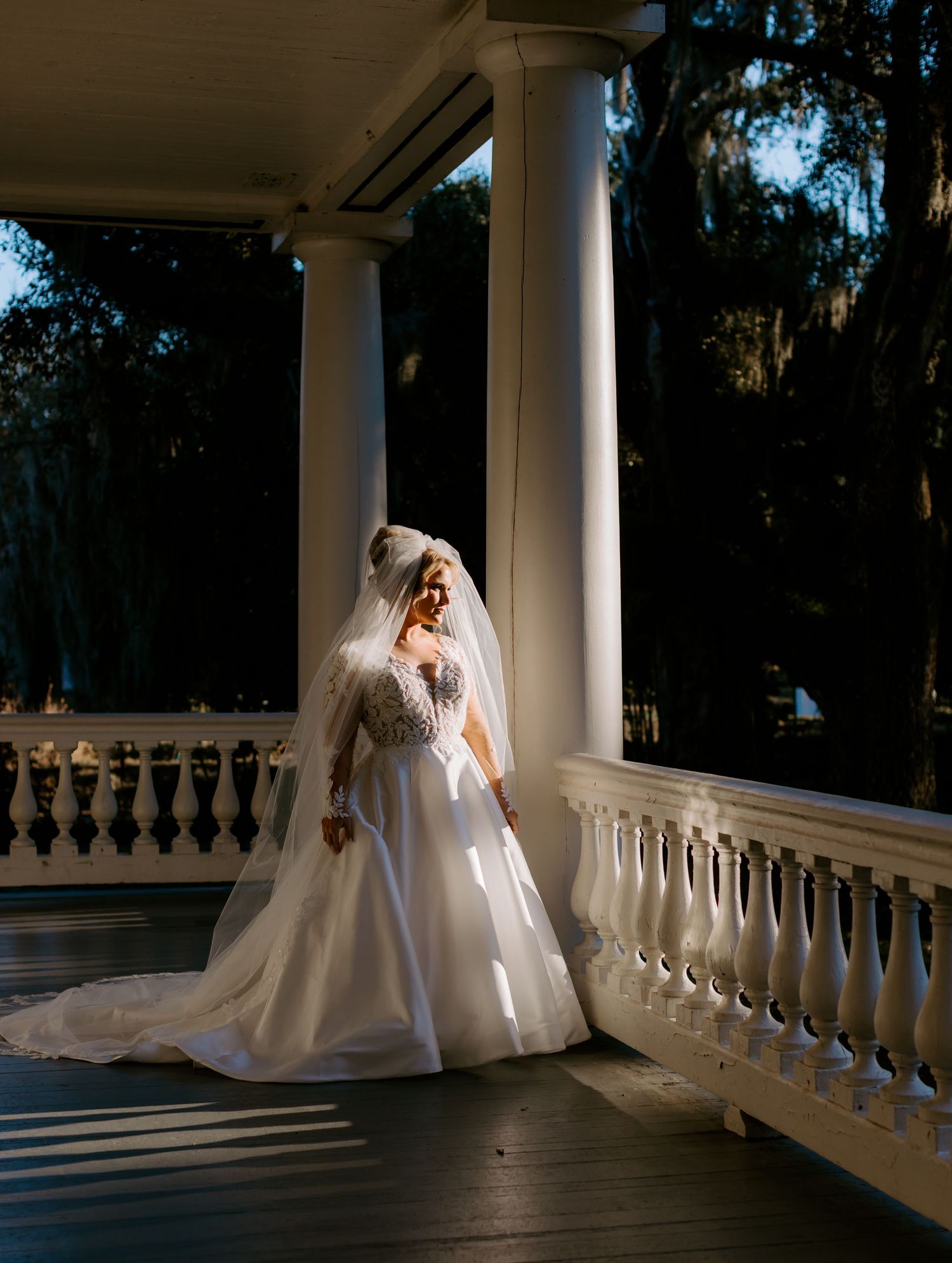 A bride in a flowing white gown and veil poses elegantly on a columned veranda with dramatic lighting and shadows.