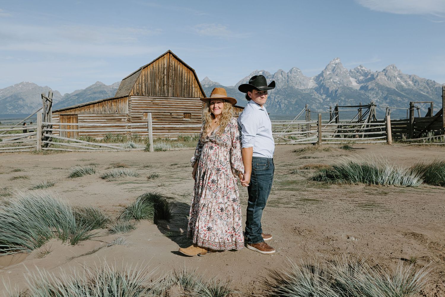 Couple holding hands in front of historic Mormon Row barn with Grand Teton mountains backdrop in Wyoming.
