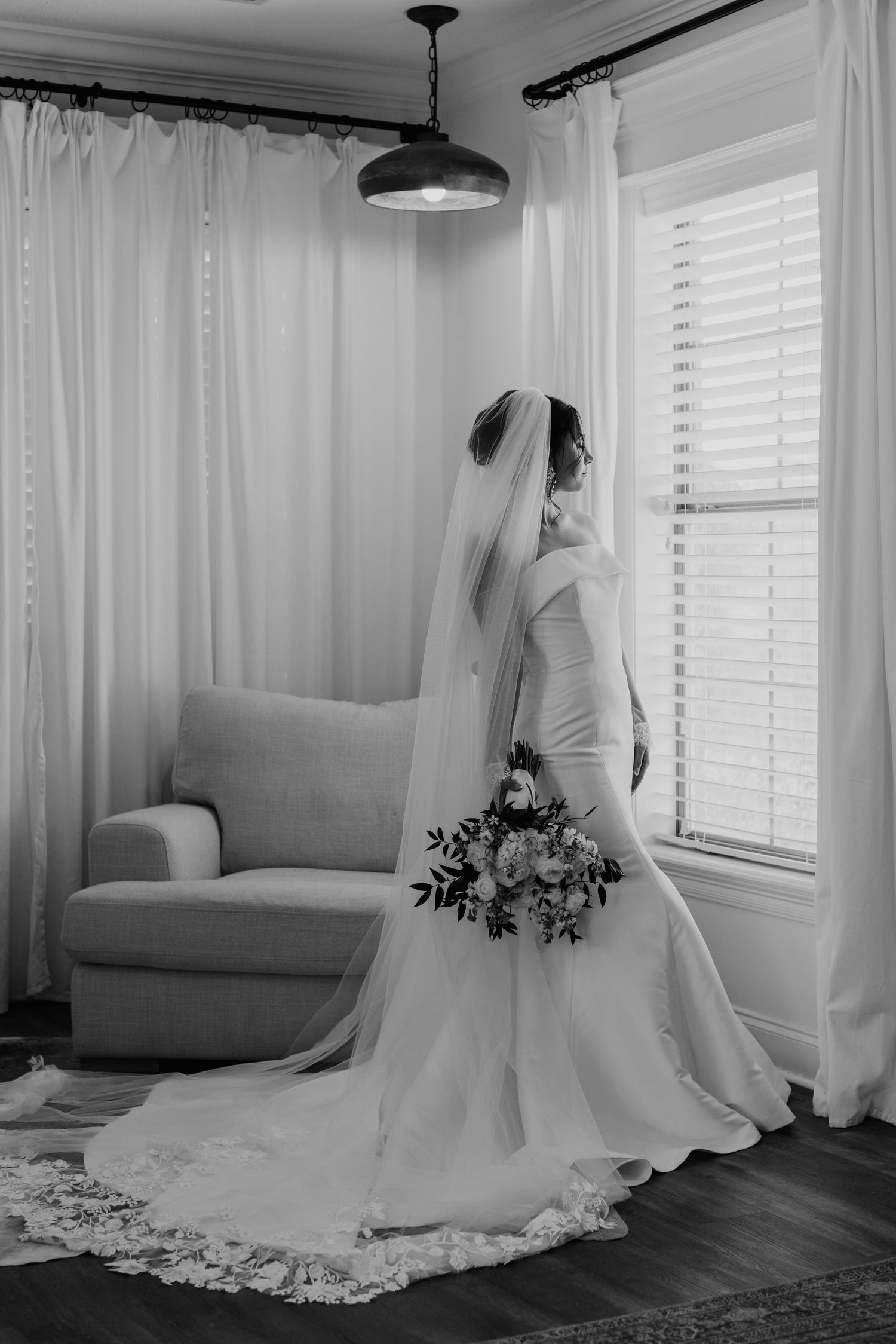 A bride in a flowing white wedding gown with lace train holds a bouquet while standing by a window in black and white.