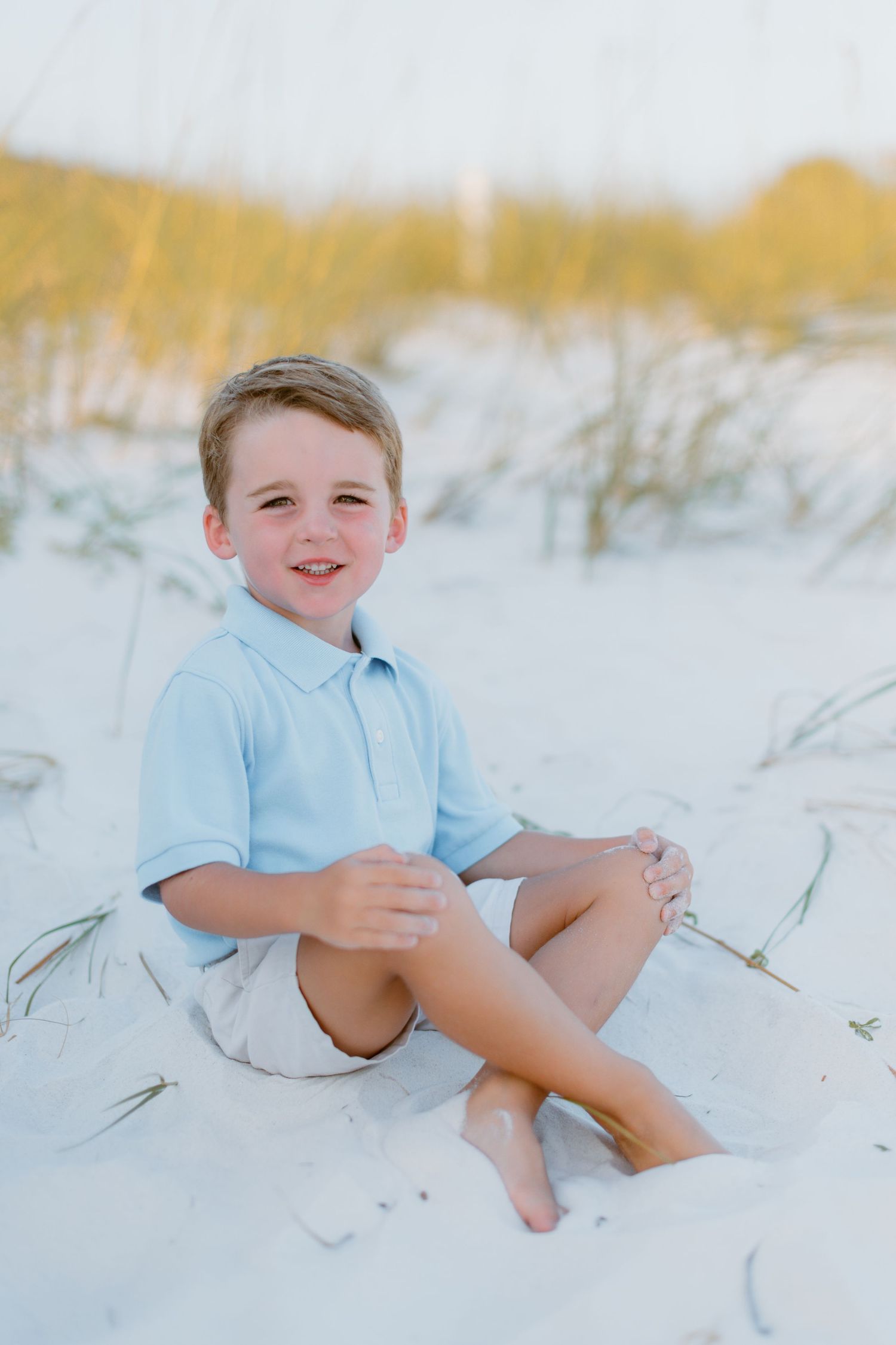 Child in light blue polo shirt playing on white sandy beach dunes during golden hour.