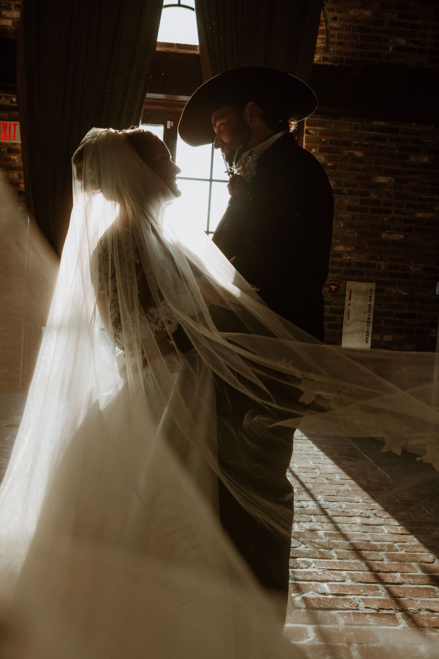 Romantic silhouette of a bride and groom dancing in front of sunlit windows with flowing veil creating dreamy atmosphere.