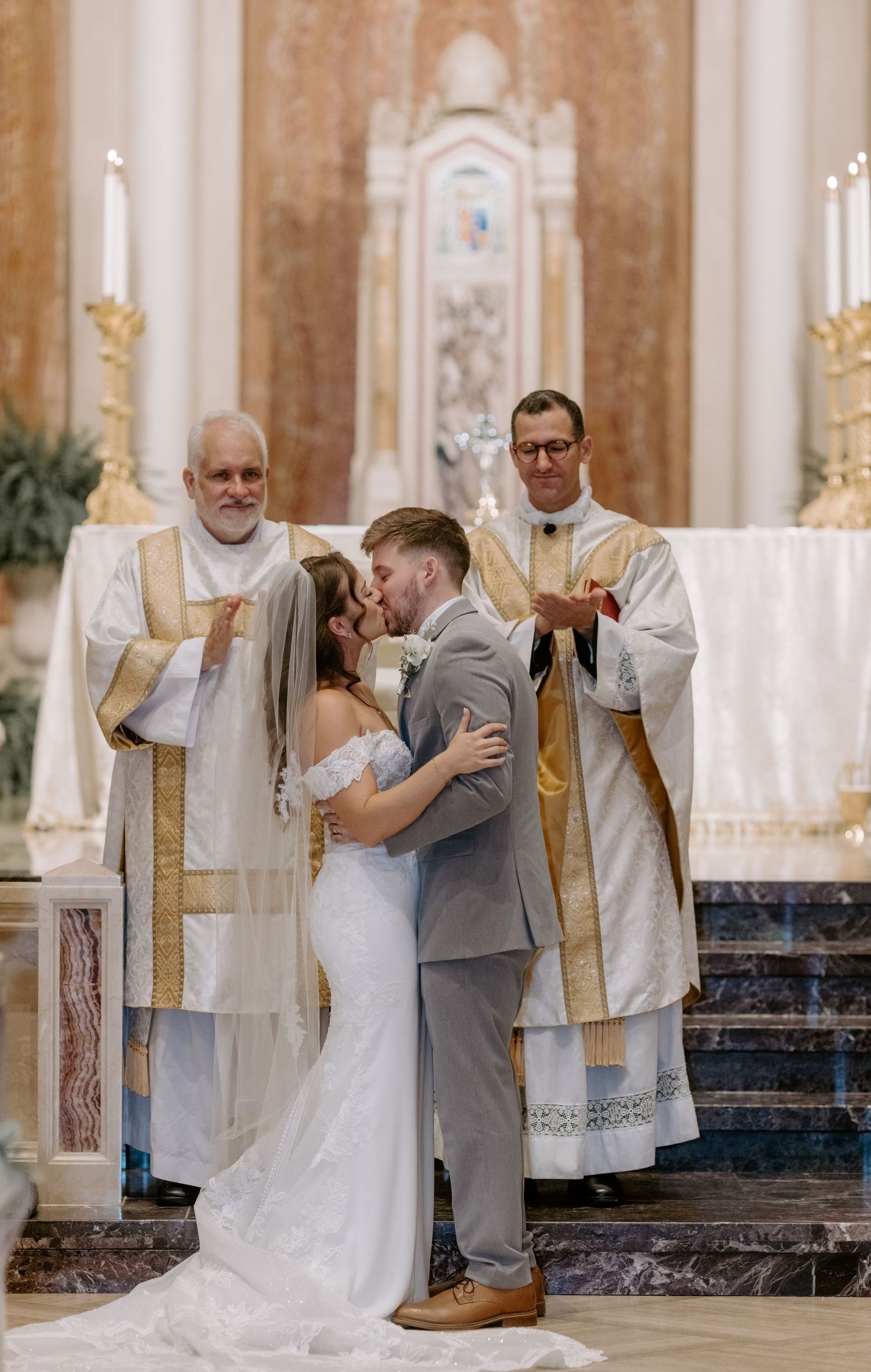 Bride and groom share first kiss at ornate church altar, flanked by two priests in gold vestments.
