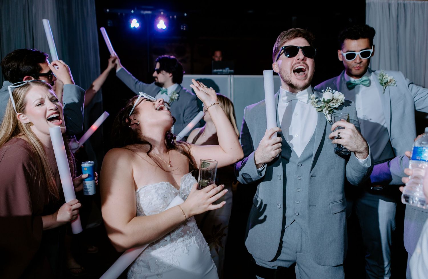 Bride and groom dancing with wedding guests holding glow sticks and wearing fun sunglasses at a lively reception party.